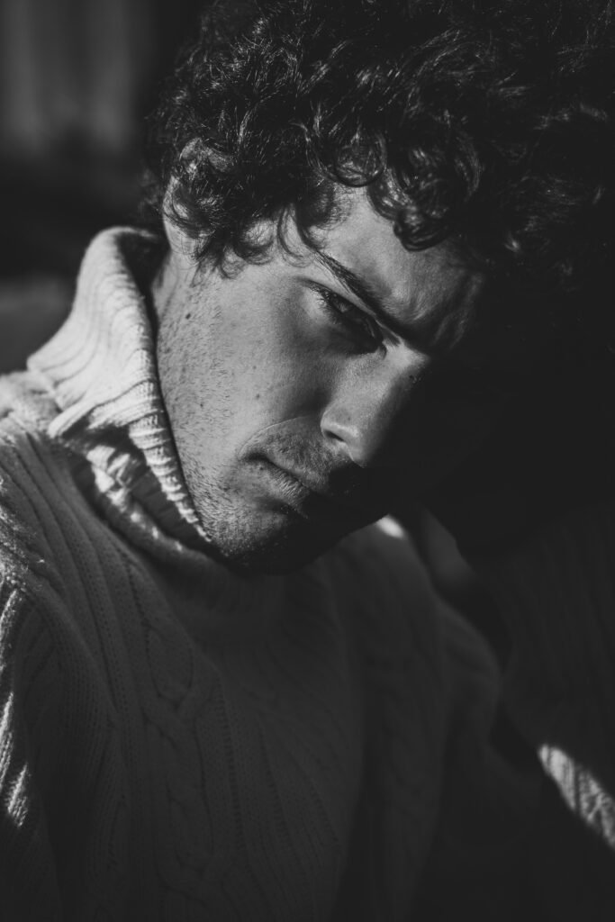 Black-and-white model portrait of a young man with curly hair in a chunky knit turtleneck, eyes lowered in soft side light