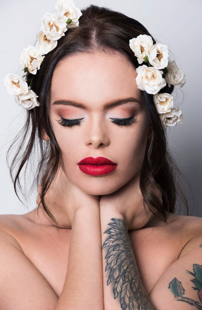 Beauty portrait of a woman with white roses in her hair, red lips and closed eyes, her tattooed arm under her chin