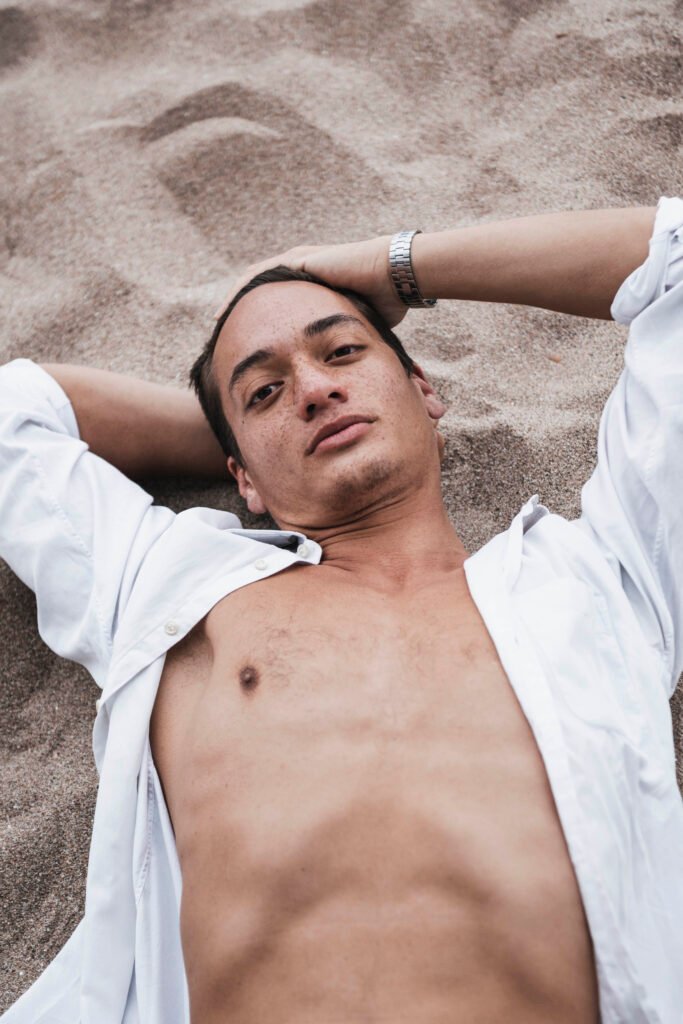 A young man lying on the sand in an open white shirt, his arms folded behind his head