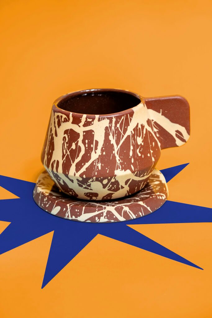 Packshot of a handmade ceramic cup and saucer with a brown and cream marbled glaze, on orange with a blue star shape