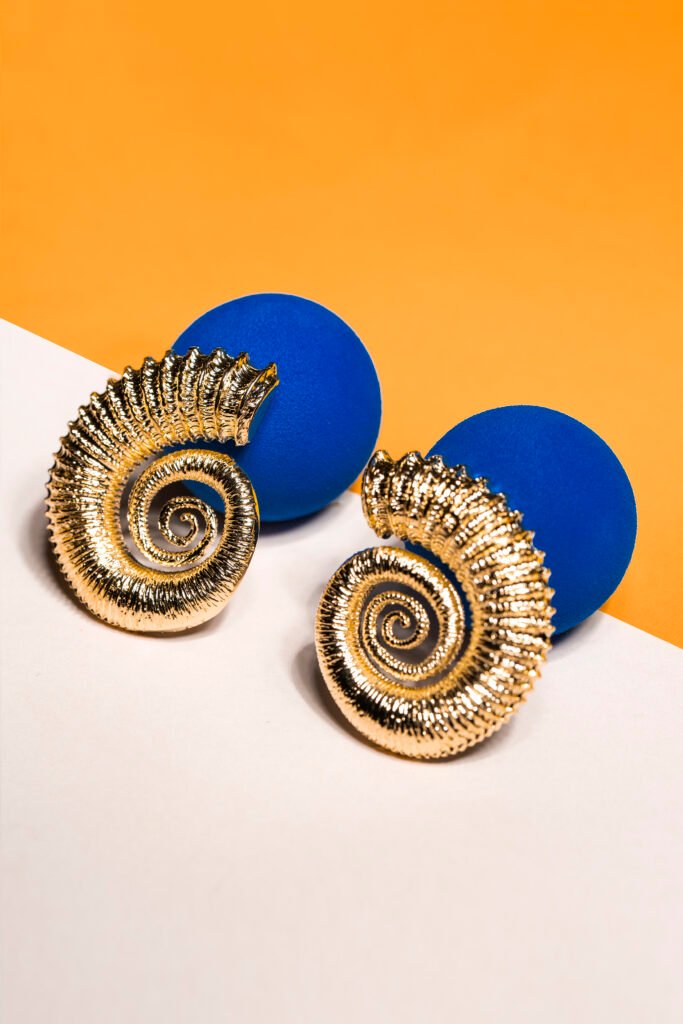 Packshot of gold ammonite-shaped earrings on two blue discs, against an orange and white background