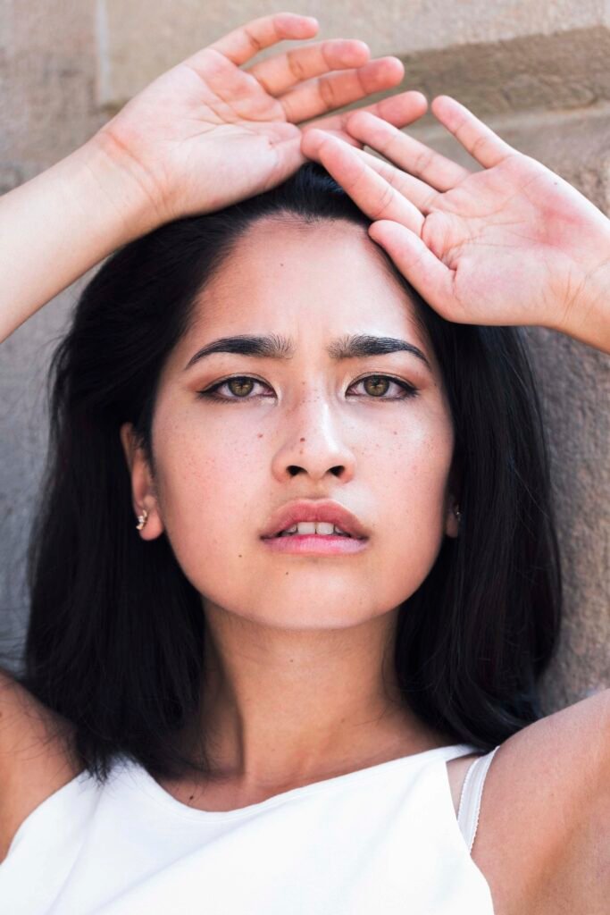Close portrait of a young woman with a black bob, her hands resting on her head, in bright sunlight
