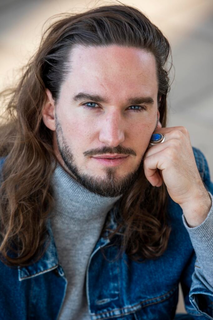 Actor portrait of a man with long wavy hair and a beard, in a denim jacket and grey turtleneck, his hand by his face