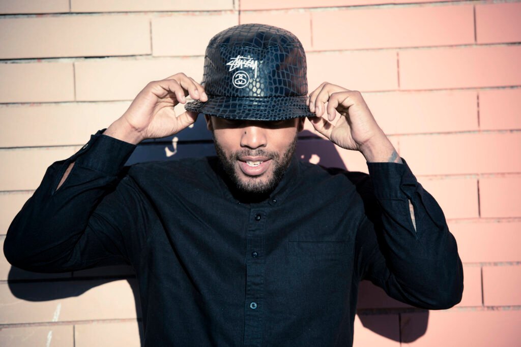 A man adjusting a black crocodile-texture Stüssy bucket hat against a pink-lit brick wall