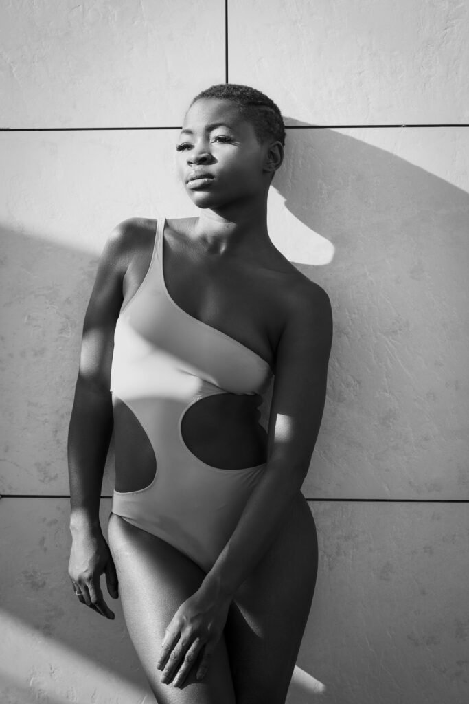 Black-and-white photo of a model in a cut-out swimsuit leaning against a concrete wall in hard sunlight