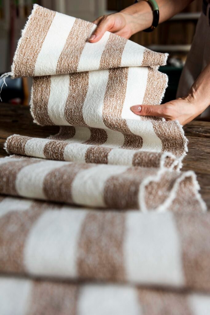 Hands unfolding handwoven textiles with brown and cream stripes in a weaving workshop