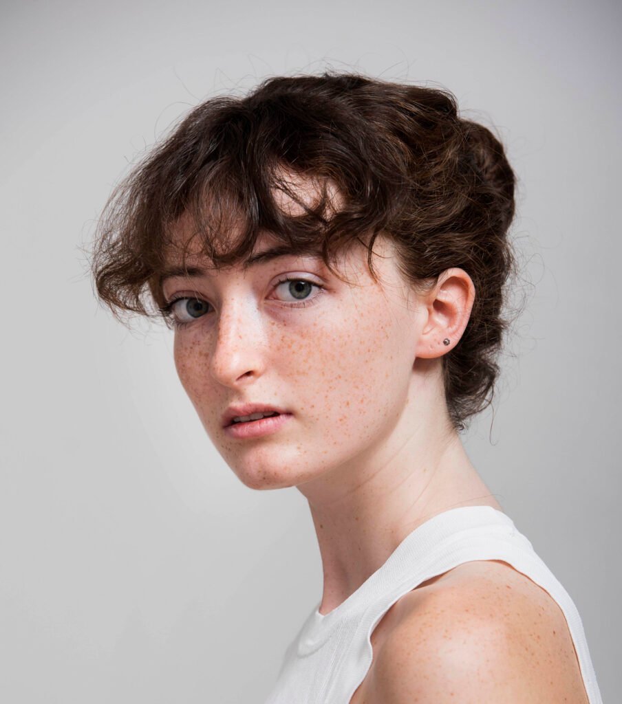 Headshot of a young woman with short curly auburn hair and freckles, in a white top against a grey backdrop