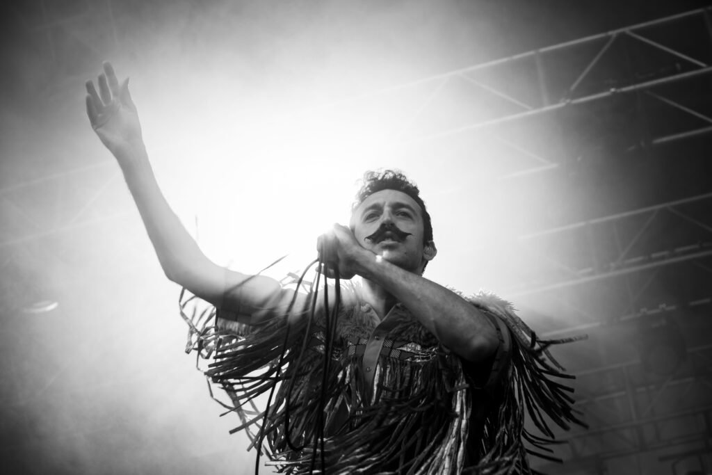 Black-and-white concert photo of a moustached singer in a fringed top raising his arm into the stage lights