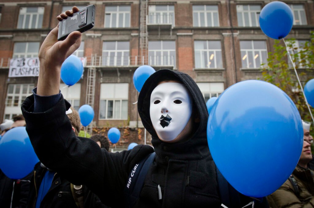 A student in a white mask with a taped mouth filming with his phone among blue balloons at a 2011 protest against austerity