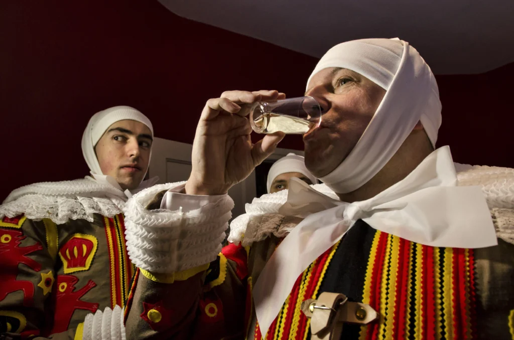 Gilles in traditional costume and white headdress at the Carnival of Binche, one of them drinking from a small glass
