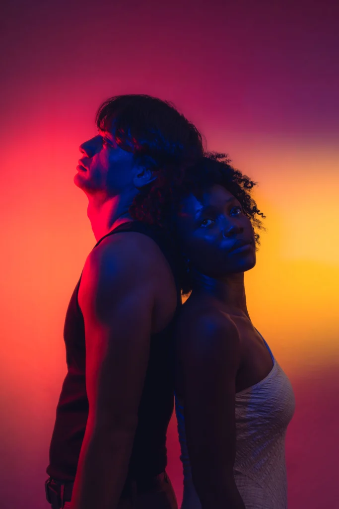 Bruin Chan and Chrystele Alix back to back, faces turned up, in red and amber light, for the play In Other Words