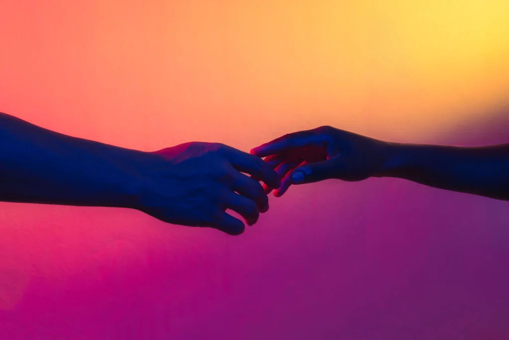 Two hands reaching for each other in red, pink and violet light, from the In Other Words shoot