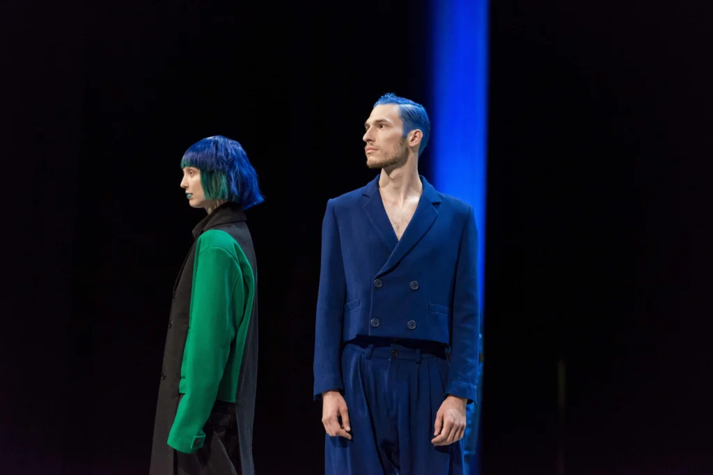 Two performers with blue hair, one in a green and black coat, one in a blue suit, on a dark stage cut by a beam of blue light