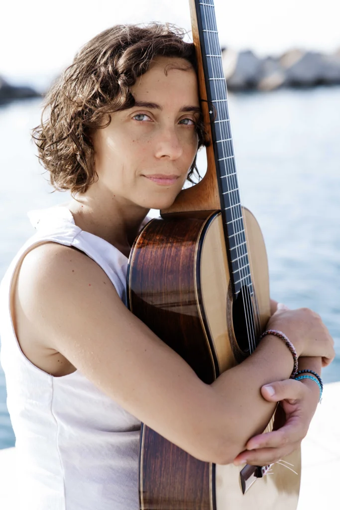 A musician with short curly hair hugging her guitar by the sea