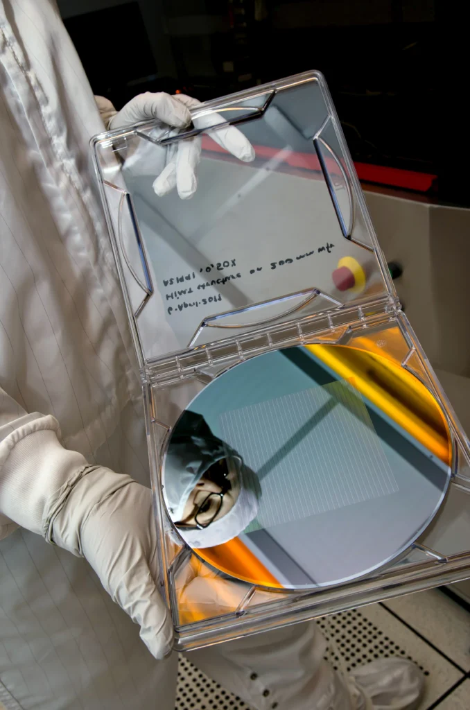 A researcher in a cleanroom suit holding up a mirror-polished silicon wafer in its clear case